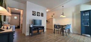 A modern living room with a flat-screen TV, a dining table with chairs, a kitchen area, and a blue cabinet.