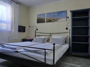 Ein ordentlich bezogenes Bett mit weißen Laken, ein Nachttisch mit Lampe und ein an der Wand befestigter Fernseher.