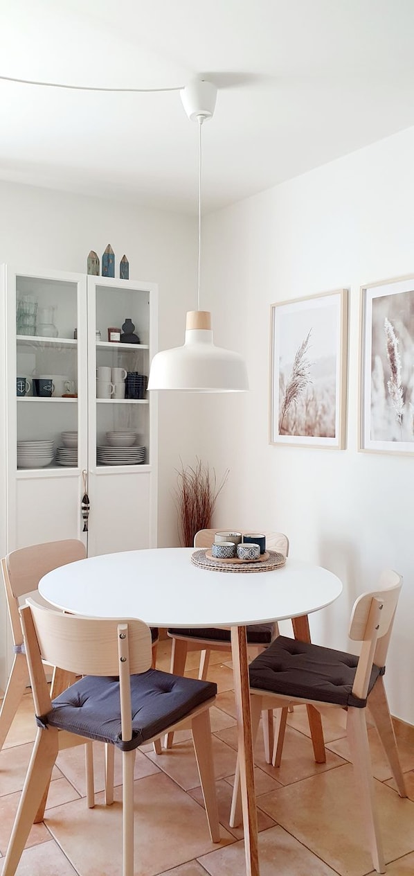 A dining area with a round white table, four chairs, a hanging pendant light, and a cabinet with glass doors.