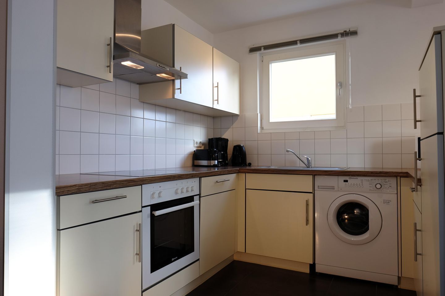 Een compacte keuken met witte kastjes, een inbouwoven, een wasmachine en een raam boven de gootsteen.