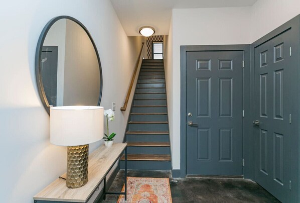 Un couloir avec un escalier en bois, une porte sombre, un miroir rond, une console en bois surmontée d’une lampe, et un tapis à motifs.