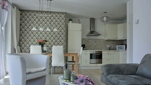 A modern living room with a grey sofa, a white armchair, and a wooden coffee table with decorative items. A kitchen with white cabinets and a microwave is visible in the background.