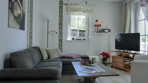 A living room with a grey sofa, a coffee table with magazines, a TV on a wooden stand, and a window with a view of greenery.
