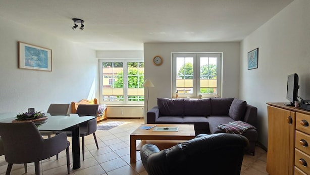 Una sala de estar con sofá, mesa de centro y mueble para la televisión. Hay un comedor con sillas y una mesa. La habitación tiene ventanas grandes y vistas a zonas verdes.