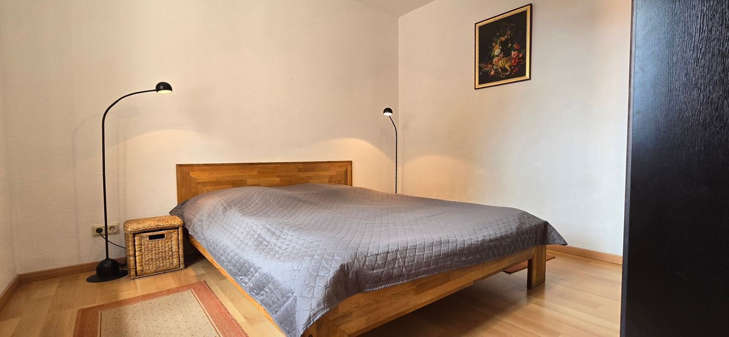 Un dormitorio con una cama de madera, dos lámparas de noche, una cesta de mimbre y un cuadro en la pared.