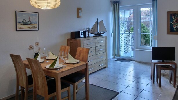A dining area with a wooden table, chairs, and a ship model on a cabinet. There is a television on a stand and a window with curtains. - Apartment O2 - 2-room apartment for 3 pers. on the ground floor with south-west facing terrace (Graal-Müritz)
