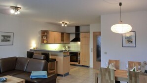 A modern living room with a sofa, a dining table, and a kitchen area with wooden cabinets and a central island. - Apartment O2 - 2-room apartment for 3 pers. on the ground floor with south-west facing terrace (Graal-Müritz)