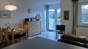 A dining area with a wooden table and chairs, a cabinet with drawers, a television on a stand, and a window with curtains. - Apartment O2 - 2-room apartment for 3 pers. on the ground floor with south-west facing terrace (Graal-Müritz)