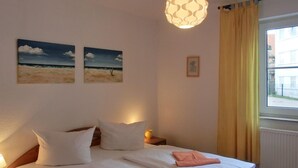 A bedroom with a bed, two paintings, a hanging light fixture, and a window with curtains. - Apartment O2 - 2-room apartment for 3 pers. on the ground floor with south-west facing terrace (Graal-Müritz)
