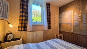 Una camera da letto con un letto, una finestra con tende, un termosifone, un comodino con una sveglia e vista sulla spiaggia.