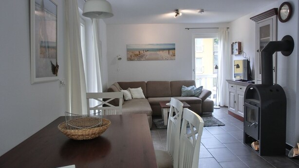 A cozy living room with a brown sofa, white chairs, a wooden table with a basket, a black stove, and a painting on the wall.