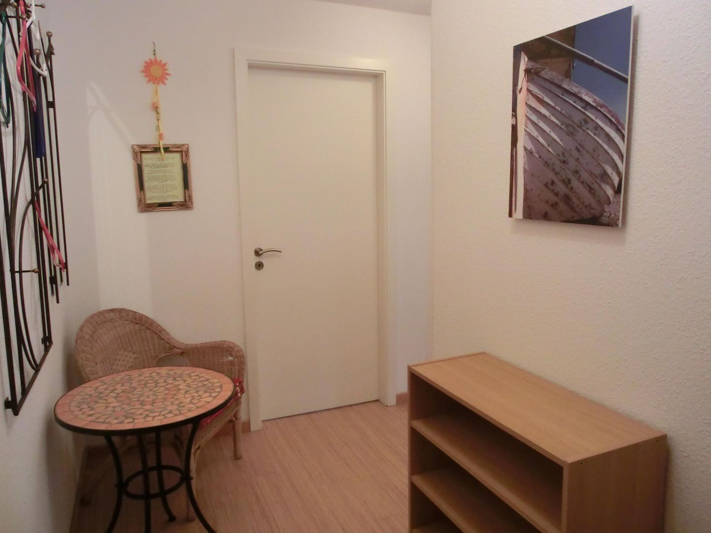 Um quarto pequeno com porta branca, uma cadeira de vime, uma mesa redonda com tampo de mosaico e uma prateleira de madeira.