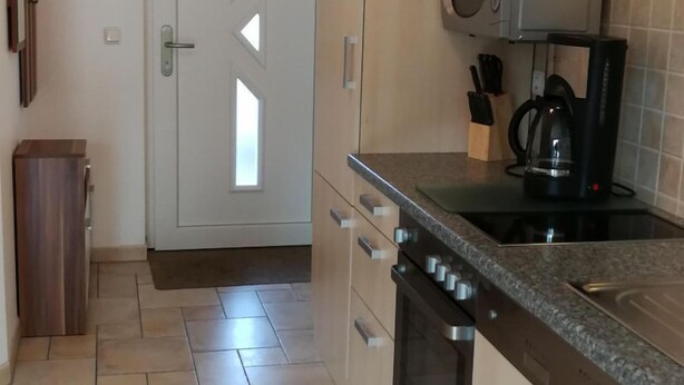 Una cocina con microondas, estufa y lavavajillas. Se observa una puerta con ventana y un cubo de basura.