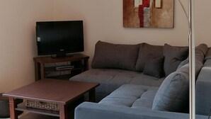 Una sala de estar con un sofá gris, una mesa de centro de madera, un televisor sobre un mueble bajo y una lámpara de pie.