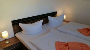 Habitación de hotel con cama doble, dos almohadas, mesita de noche con lámpara y una toalla naranja sobre la cama.