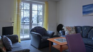 A living room with a sofa, armchair, and a small table. There is a window with curtains, a radiator, and a painting on the wall. - Apartment i25 - 56 m² large 3-room apartment 3 pers. on the 1st floor in the house Lee (Graal-Müritz)