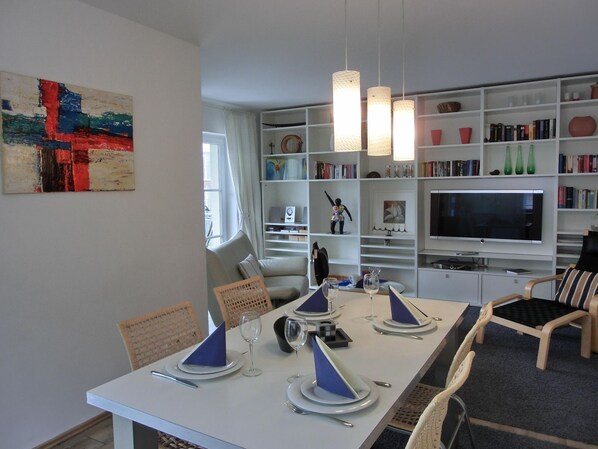 A modern living room with a dining table set for four, a bookshelf filled with books and decorative items, and a large flat-screen TV.