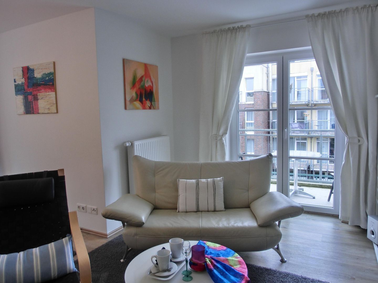 A living room with a beige sofa, a coffee table with a tray, a black chair, a radiator, and a window with curtains.