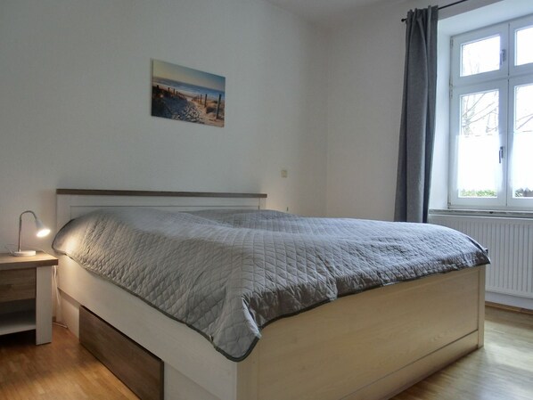 Une chambre à coucher avec un lit, une table de chevet avec une lampe, une fenêtre avec des rideaux et un tableau au mur.
