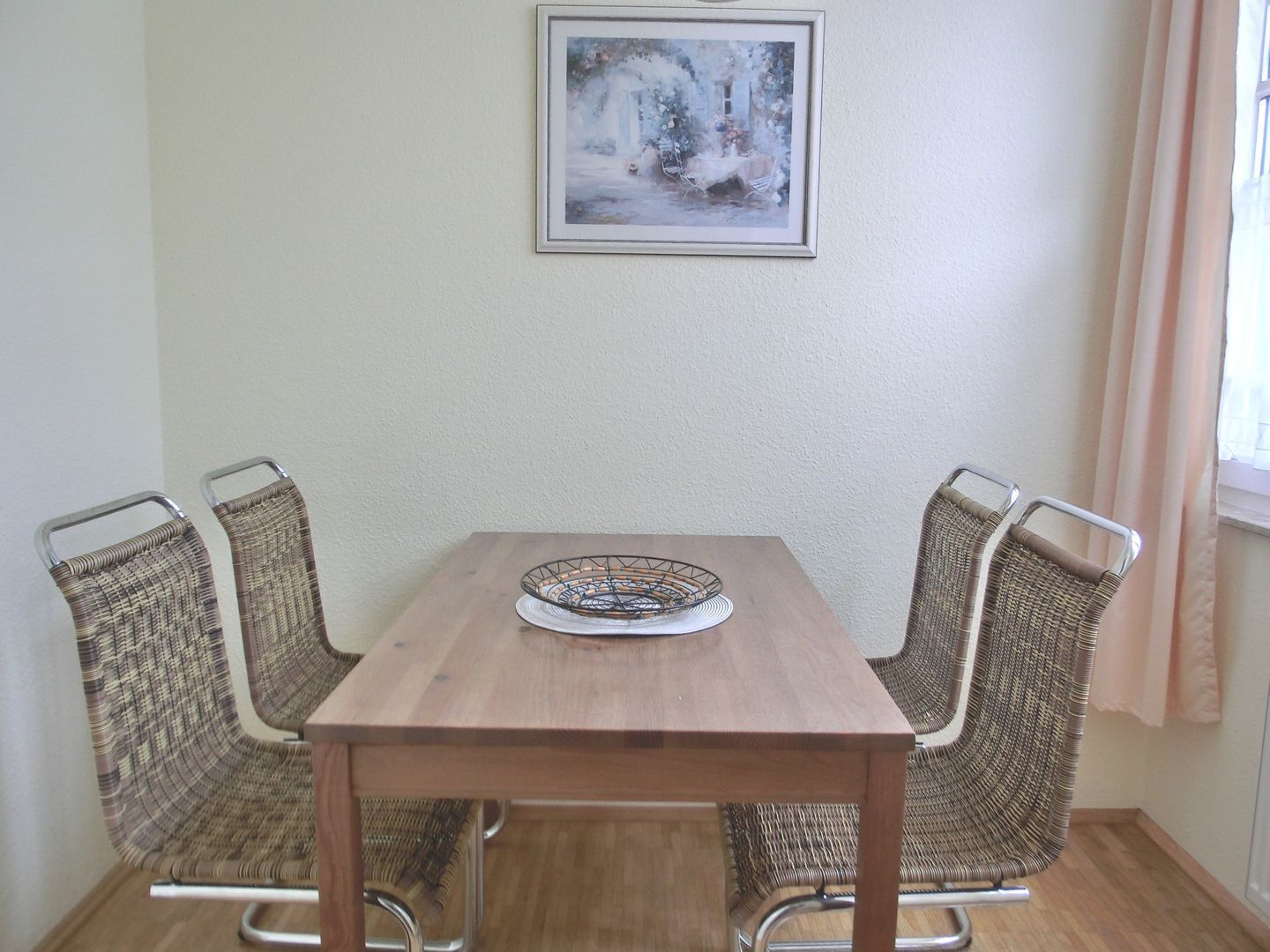 Un tavolo da pranzo con una sedia intrecciata su ogni lato, un quadro incorniciato sopra e una finestra con tende.