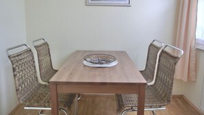 A dining table with a woven chair on each side, a framed painting above, and a window with curtains. - Vacation apartment LVC11 - 2-room apartment for 4 pers. with WLAN, pets allowed (Graal-Müritz)