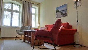 A modern living room with a red sofa, a grey armchair, and a wooden coffee table. There is a large window with curtains, a floor lamp, and a painting on the wall. - Vacation apartment LVC11 - 2-room apartment for 4 pers. with WLAN, pets allowed (Graal-Müritz)