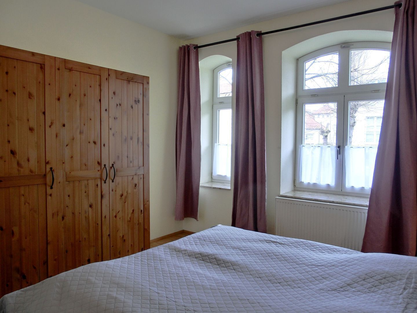 A bedroom with a bed, wooden wardrobe, and windows with curtains.