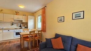 Ett kompakt kök med vita skåp, ett matbord och stolar. I vardagsrummet finns en blå soffa med orange kuddar.