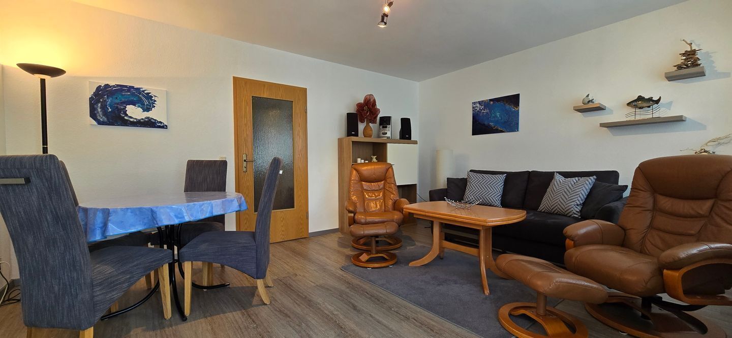 A modern living room with a dining table, chairs, a sofa, and a recliner. There are wall-mounted shelves with decorative items.