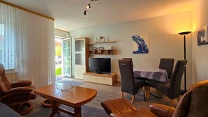 A living room with a TV, a dining table, and chairs. There is a window with curtains, a sofa, and a lamp. - Vacation apartment P1 - 2-room apartment with terrace in the town center (Graal-Müritz)