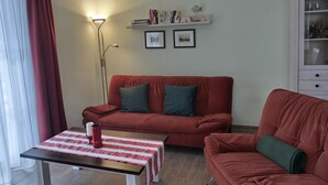 A living room with a red sofa, a table with a red and white tablecloth, and a floor lamp. There are bookshelves with books and framed pictures on the wall. - Apartment L32 - "Lütte Möw" - 2-room apartment for 4 people on the 1st floor with WiFi (Graal-Müritz)