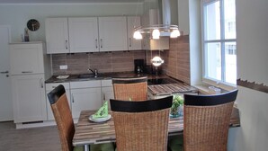 A modern kitchen with white cabinets, a dark countertop, and a large window. There is a dining table with wicker chairs and a clock on the wall. - Apartment L32 - "Lütte Möw" - 2-room apartment for 4 people on the 1st floor with WiFi (Graal-Müritz)