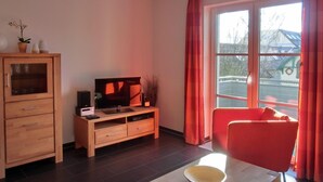 A modern living room with a wooden cabinet, a flat-screen TV, a red chair, and a large window with curtains. - Vacation apartment L31- "Fürstenhof" - 2-room apartment for 4 people on the 2nd floor with WLAN (Graal-Müritz)