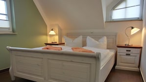 Et soveværelse med en hvid seng, to sengeborde med lamper, et trænatbord og et vindue med persienner.
