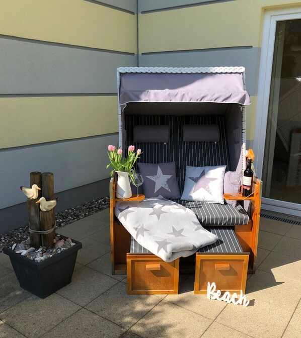 Un espace extérieur aménagé avec des sièges en bois, surmonté d’un auvent, agrémenté de coussins à motifs étoilés, d’un vase rempli de fleurs et d’une bouteille de vin.