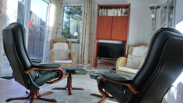 Uma sala de estar com duas poltronas de couro preto, um sofá de vime, uma televisão sobre um suporte de madeira e uma estante repleta de livros.