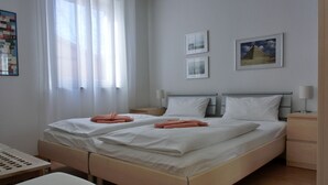 Cama de casal com lençóis brancos e toalhas laranja, duas mesinhas de cabeceira com abajures, um banco de madeira e uma janela com cortinas finas.