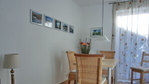 Uma área de refeições com cadeiras de vime, uma mesa e um abajur. Há quadros nas paredes e uma janela com cortinas florais.
