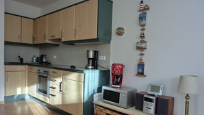Cozinha compacta com micro-ondas, cafeteira e torradeira em uma bancada. Há cestos de vime sob a bancada e um abajur à direita.