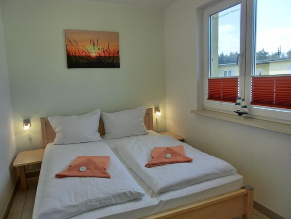 A double bed with white bedding and two orange towels, a nightstand, a wall-mounted lamp, and a window with blinds.