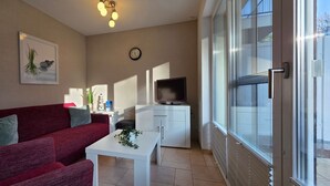 A living room with a red sofa, a white coffee table, a TV on a cabinet, and a large sliding glass door to the outdoors. - Apartment KWi 46 ground floor - 2-room apartment for 4 pers. on the ground floor, incl. 4 bicycles (Gelbensande)