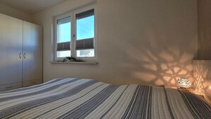 A bedroom with a bed, a wardrobe, a window with blinds, and a wall-mounted light fixture. - Apartment KWi 46 ground floor - 2-room apartment for 4 pers. on the ground floor, incl. 4 bicycles (Gelbensande)