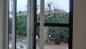 Une porte-fenêtre coulissante donne accès à un patio extérieur doté d’un sol en briques, d’une table avec une chaise et d’une poubelle portant l’inscription « Eenders ».