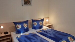 En snyggt bäddad säng med blått sänglinne och blommiga kuddar, en sänglampa och en liten hylla med en radio.
