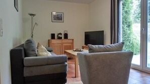 Ett modernt vardagsrum med en grå soffa, en grå fåtölj, ett träskåp med en platt-TV och ett stort fönster med utsikt över grönområden.