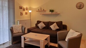 Una sala de estar moderna con un sofá marrón, dos sillones, una mesa de centro y una lámpara de pie. Se observan estantes empotrados con objetos decorativos y un reloj.
