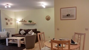 Una sala de estar con sofá, mesa de centro y mesa de comedor con sillas. Hay un reloj en la pared y un cuadro enmarcado sobre la mesa de comedor.