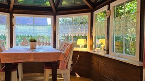 Une terrasse couverte avec une table dressée pour le repas, entourée de chaises, et de grandes fenêtres ornées de rideaux en dentelle.
