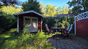 Une cabane en bois avec une porte et des fenêtres, un portail rouge et une table avec des chaises, le tout situé dans un jardin.
