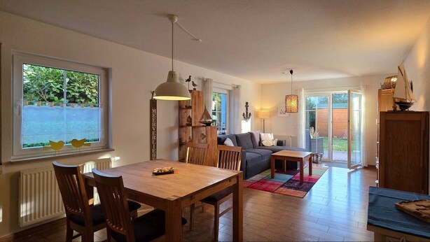 Una sala de estar acogedora con una mesa de comedor de madera, un sofá y una mesa de centro. Se ve un ventanal con vistas a la vegetación.